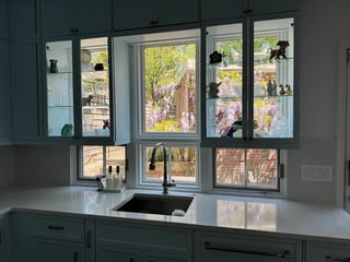 Kitchen sink with dark green cabinetry, white countertop, and large multipane windows overlooking a street lined with trees and buildings