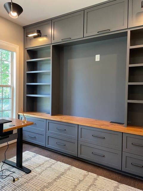 Modern home office with gray built-in shelving, wooden countertop desk, and large dark wall panel for media display