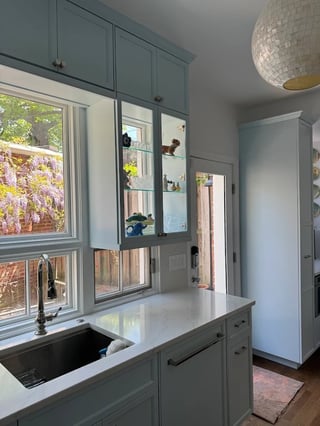 Modern kitchen with sage green cabinets, white countertop, window overlooking garden, glass-front upper cabinets, and decorative pendant light