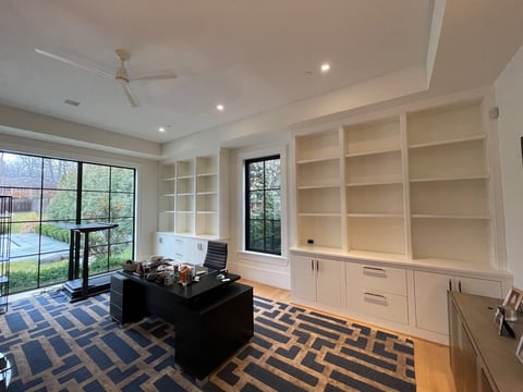 Modern home office with built-in white shelving, black desk, geometric area rug, and views of backyard through large windows