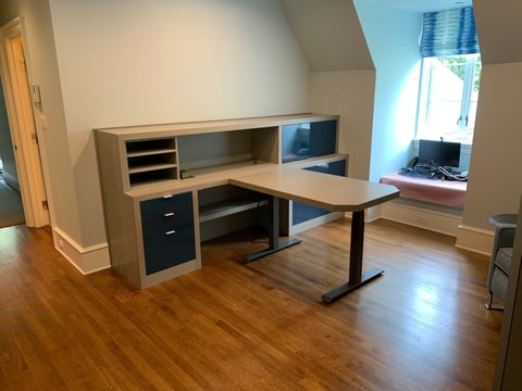 Modern home office with L-shaped desk, storage shelving, hardwood floors, and natural light from a window in an attic room